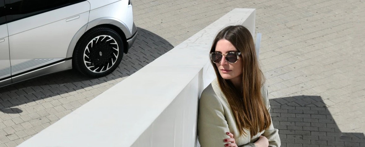 Britta Reineke sitting behind a wall in front of a gray IONIQ 5. She has long brunette hair and is wearing large tortoise shell sunglasses and a beige blazer.